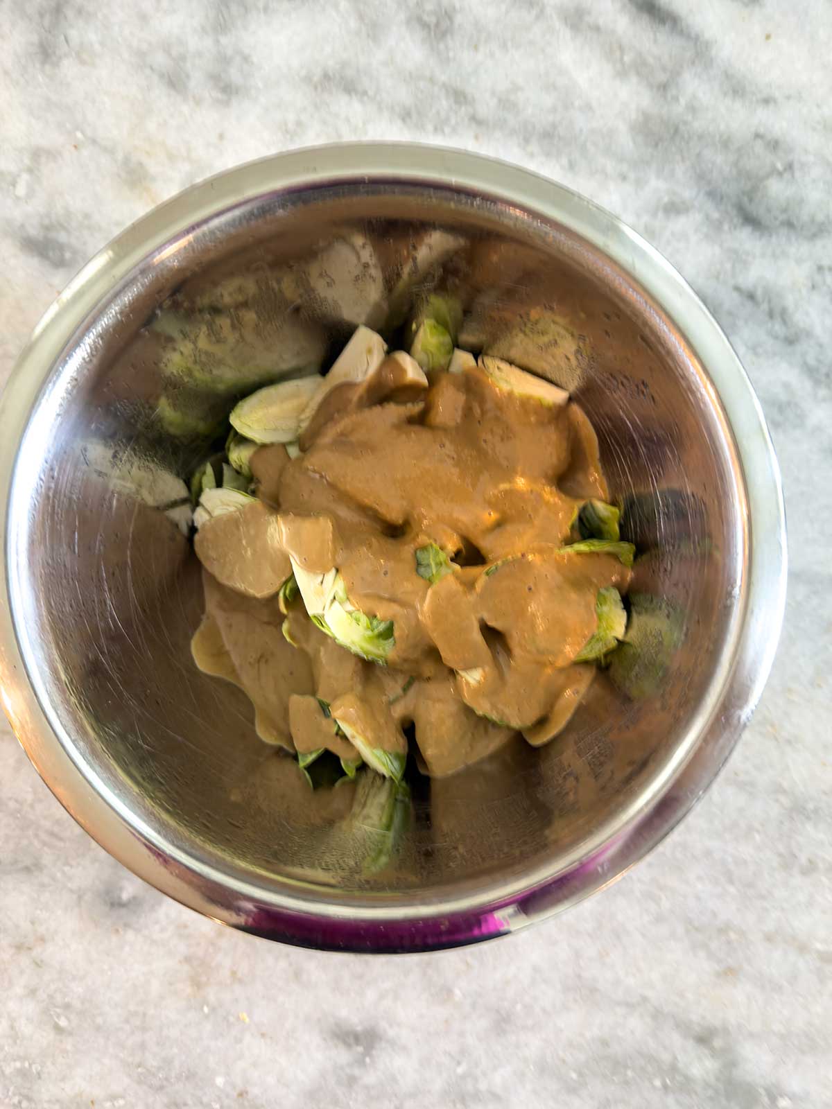 A stainless steel bowl containing chopped Brussels sprouts topped with a creamy brown sauce, placed on a light gray marble surface.