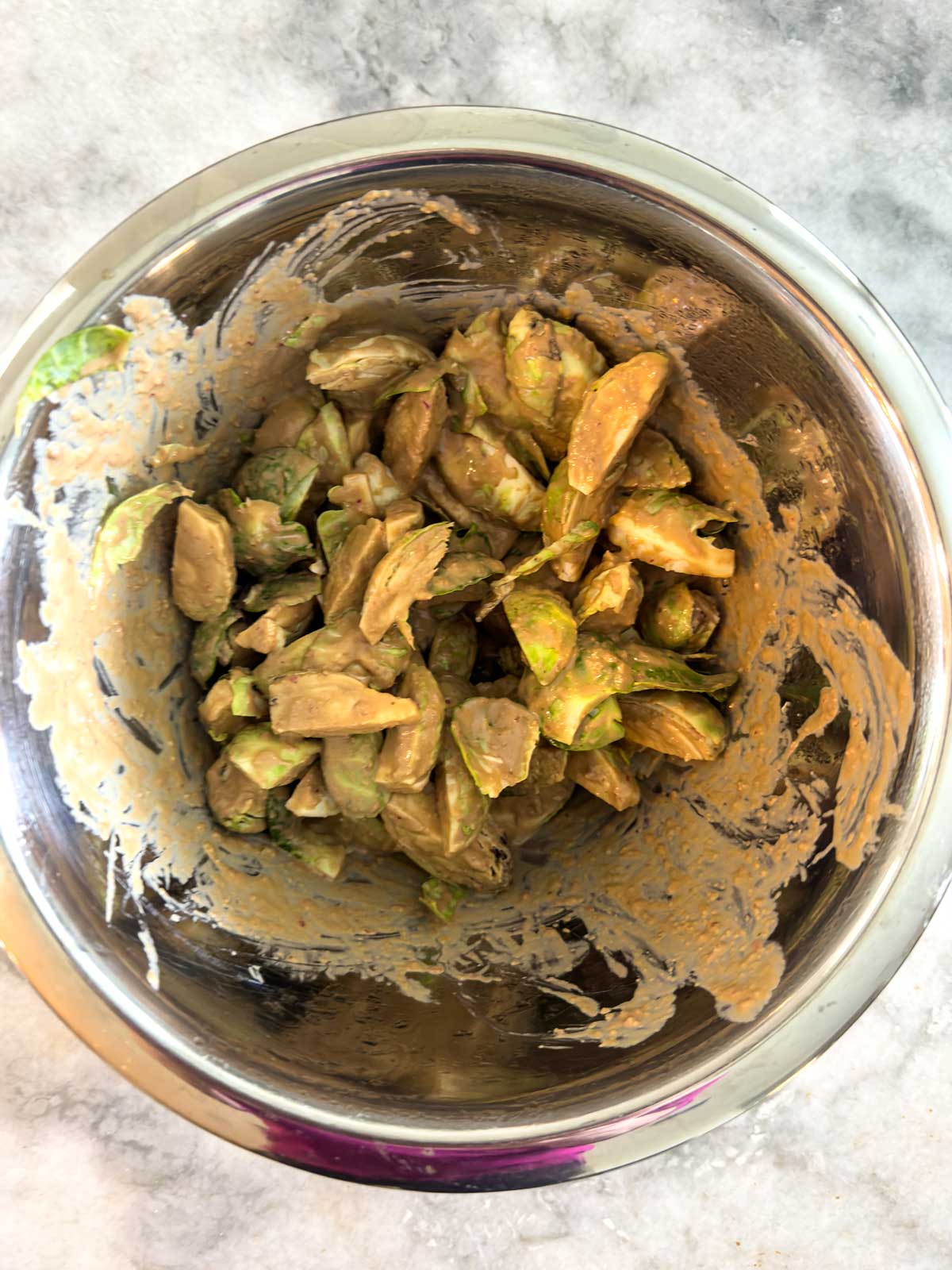 A metal mixing bowl containing sliced Brussels sprouts coated in a creamy, beige-colored dressing, placed on a light gray countertop.