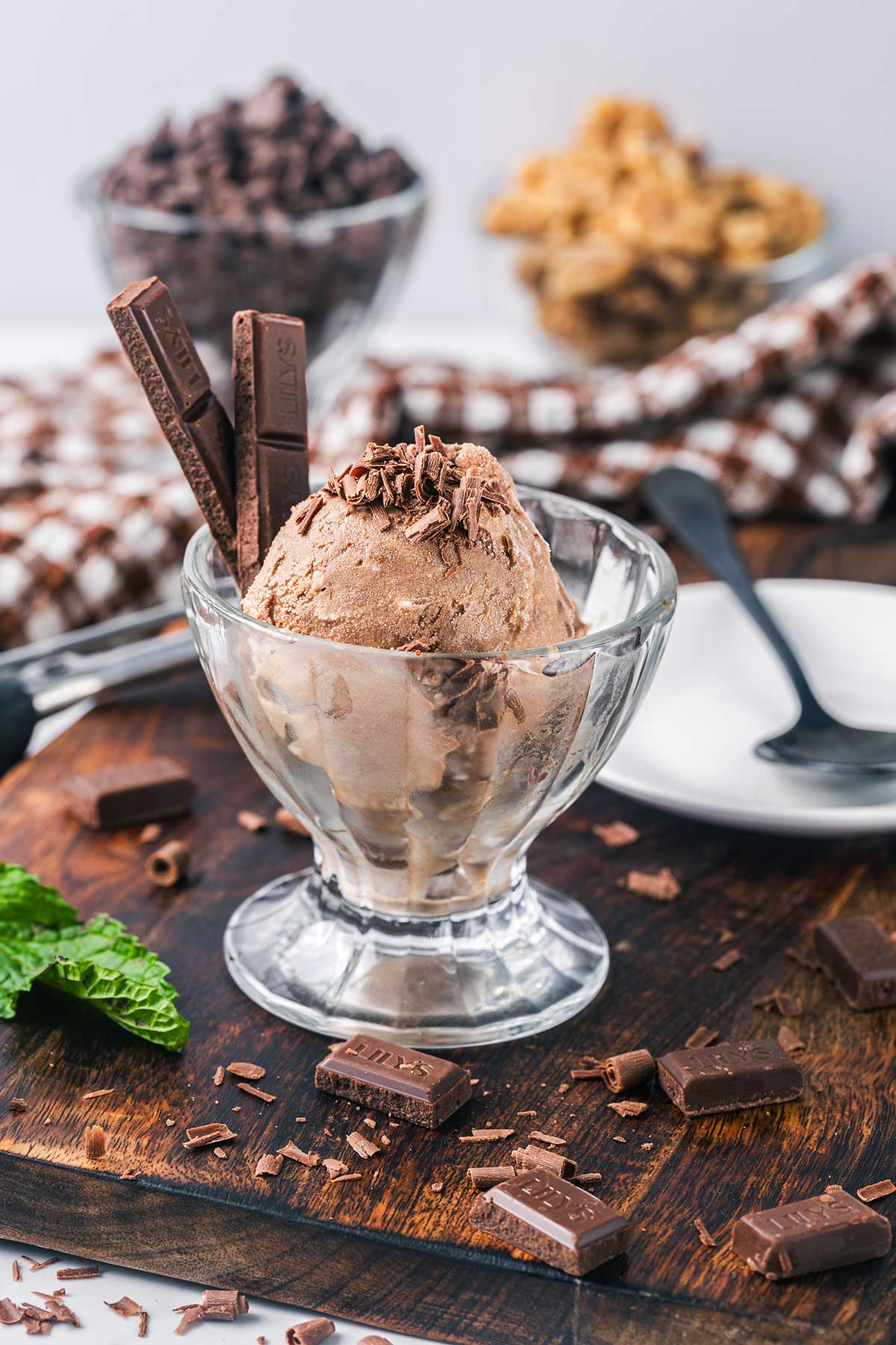 A glass bowl filled with chocolate ice cream, topped with chocolate shavings and two chocolate bars. The bowl sits on a wooden surface with chocolate pieces and a mint leaf nearby; blurred dessert and utensils in the background.