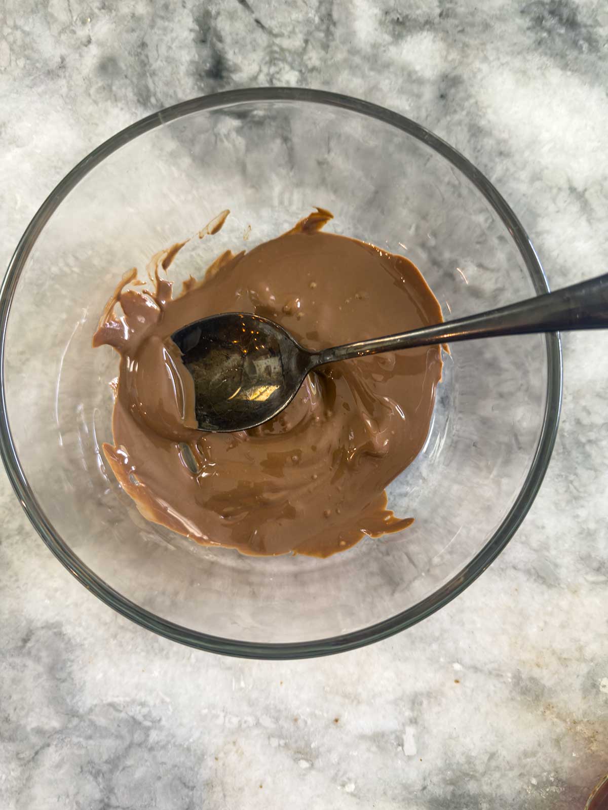 A glass bowl containing melted milk chocolate, perfect for making Ninja Creami chocolate ice cream, with a metal spoon resting inside, placed on a marble surface.