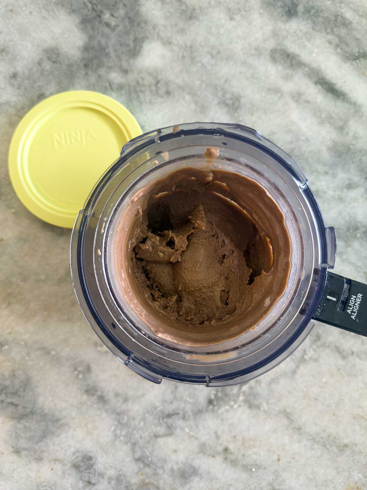 A blender cup filled with Ninja Creami chocolate ice cream sits on a marble surface, its yellow lid placed nearby.