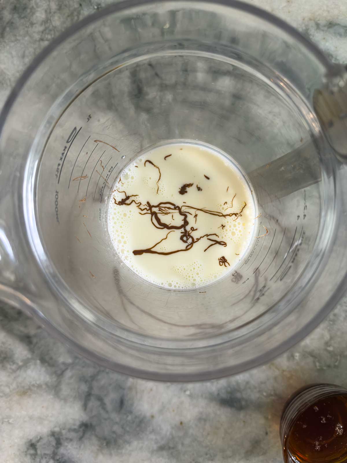 A clear measuring pitcher with milk and streaks of chocolate syrup inside, perfect for making Ninja Creami chocolate ice cream, placed on a marble countertop.
