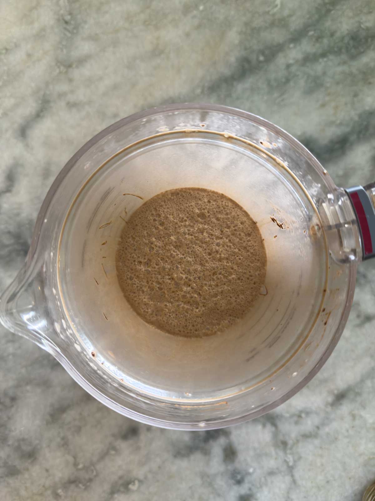 A glass measuring cup with bubbly brown liquid at the bottom, viewed from above, placed on a light gray marble surface—the start of homemade Ninja Creami chocolate ice cream.