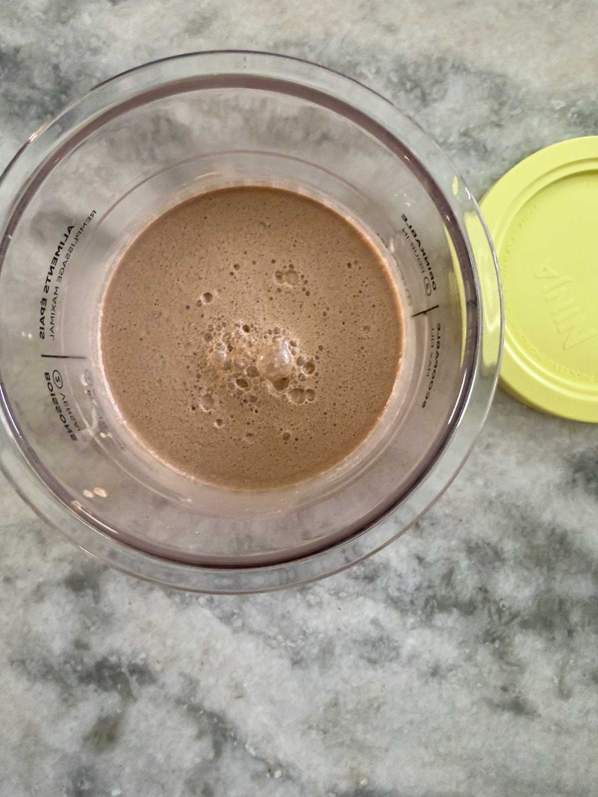 A top-down view of a transparent cup filled with frothy chocolate milk—reminiscent of ninja creami chocolate ice cream—rests on a gray marble surface, with a yellow lid beside it.