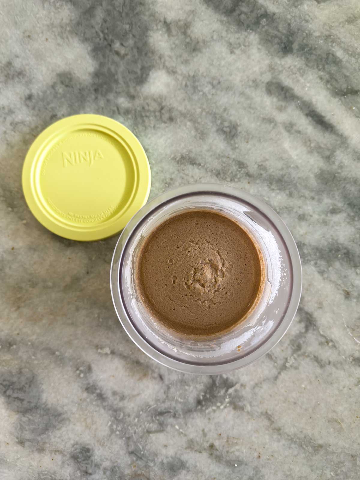 A jar with a yellow lid labeled Ninja sits open on a gray marble surface, revealing smooth, brown ninja creami chocolate ice cream inside.