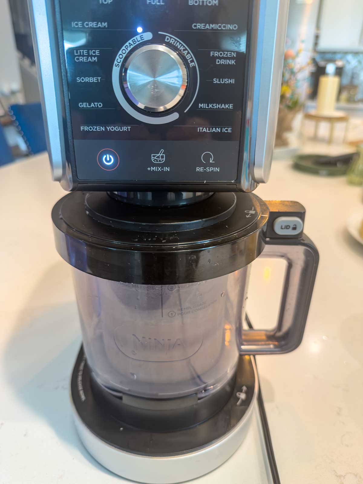 A close-up of a Ninja Creami ice cream maker on a kitchen counter, showing its control panel with various dessert options and a clear container filled with delicious chocolate ice cream attached at the base.