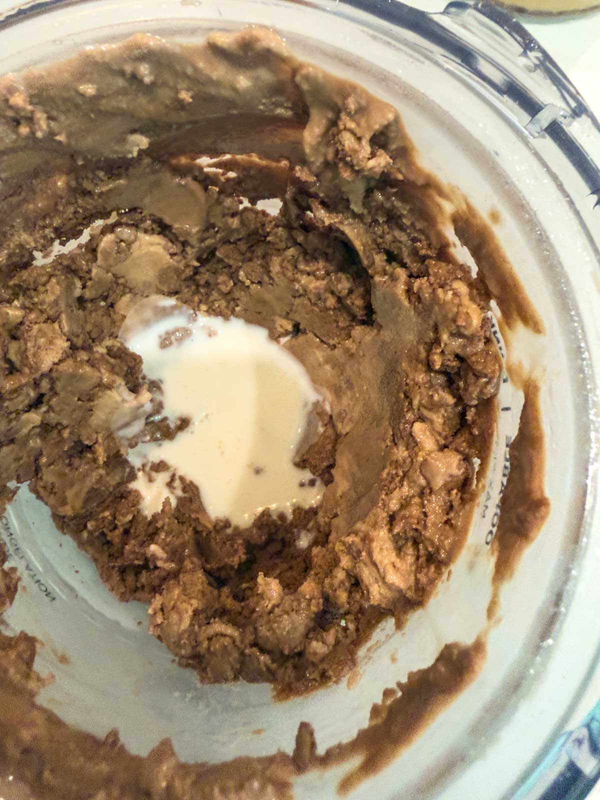 A close-up of a blender with thick ninja creami chocolate ice cream mixture stuck to the sides and a small pool of liquid, possibly milk, in the center. The mixture appears chunky and creamy.