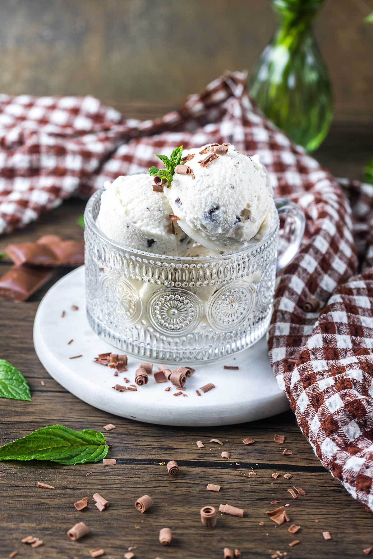 A glass cup filled with three scoops of vanilla ice cream with chocolate chips, garnished with mint and chocolate shavings, sits on a white plate with a brown checkered cloth and chocolate pieces nearby.