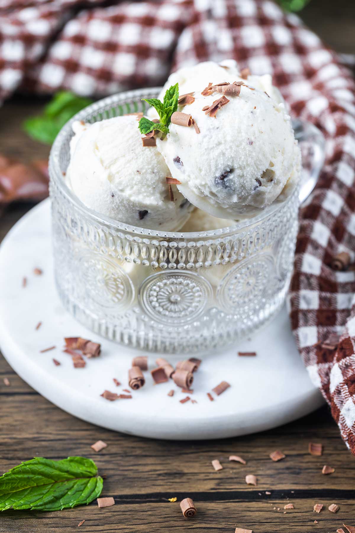 A glass cup filled with scoops of vanilla ice cream with chocolate shavings, garnished with a mint sprig, sits on a white plate. A brown and white checkered cloth and mint leaves are nearby on a wooden surface.