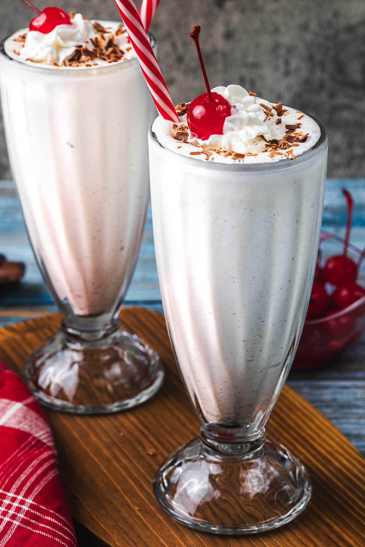 Two tall milkshake glasses filled with vanilla milkshakes, topped with whipped cream, chocolate shavings, and a cherry. One has a red striped straw. A bowl of cherries and a red napkin are in the background.