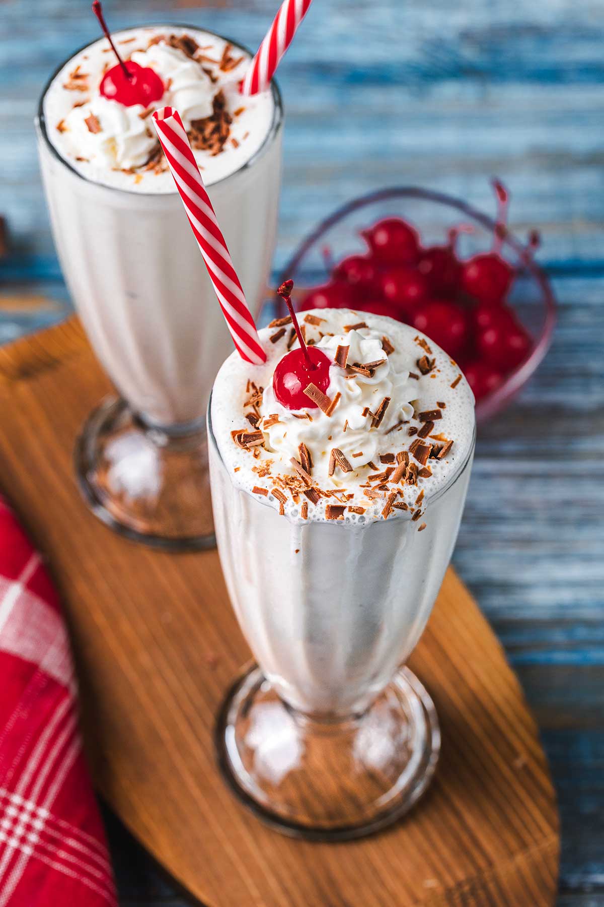 Two vanilla milkshakes topped with whipped cream, chocolate shavings, and a cherry, each with a red-striped straw, served in tall glasses on a wooden board, with maraschino cherries in a bowl nearby.
