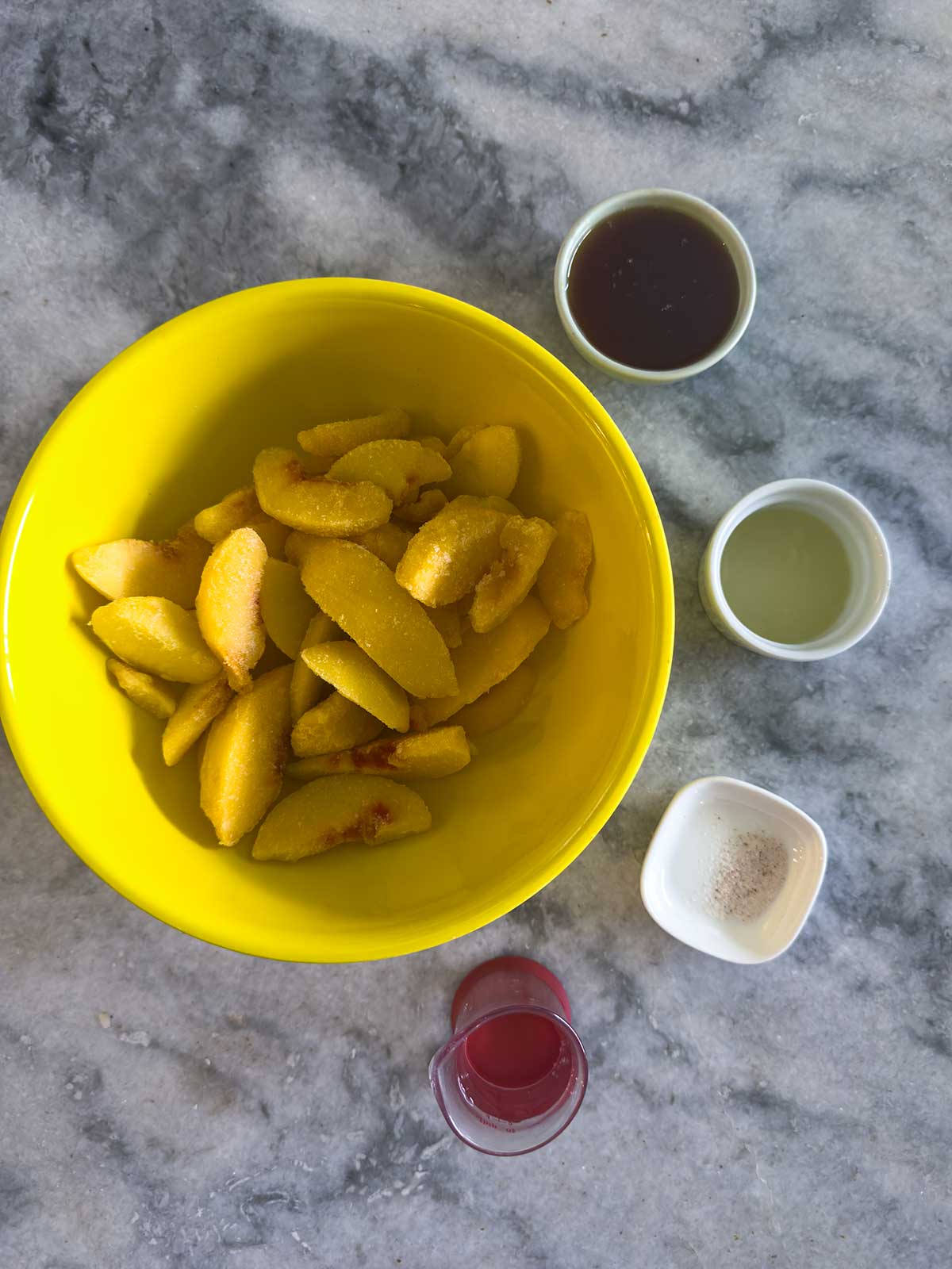 Sliced peaches in a yellow bowl on a marble surface, surrounded by small containers of brown liquid, clear liquid, reddish liquid, and a white granular substance.
