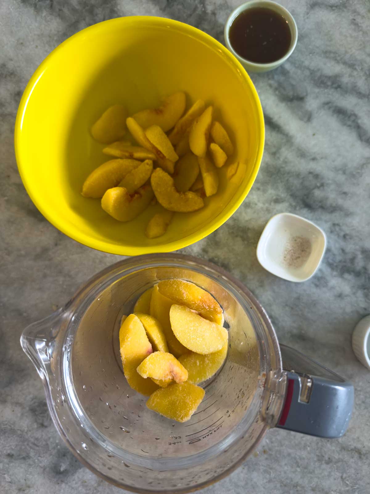 Frozen peach slices in a yellow bowl and a blender, with small bowls of brown liquid and white powder on a gray countertop, ready for making a recipe.