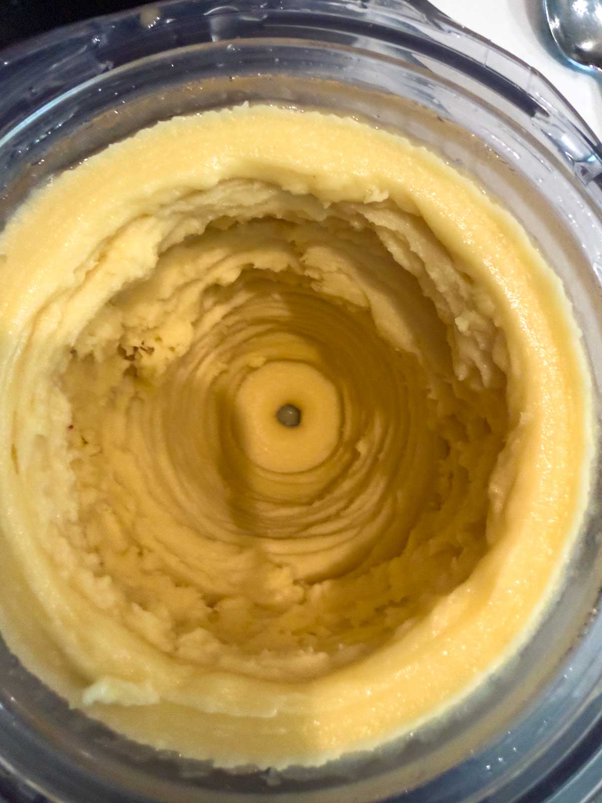 A top-down view of creamy, yellow mashed potatoes inside a mixing bowl, with a swirl pattern and a small hole in the center.