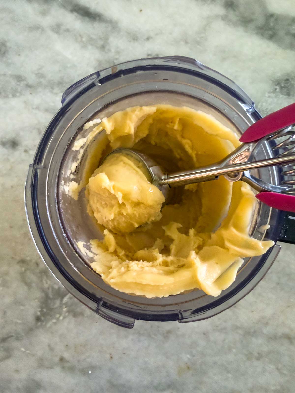 A metal ice cream scoop is being used to serve creamy, yellow ice cream from a food processor bowl on a marble countertop.