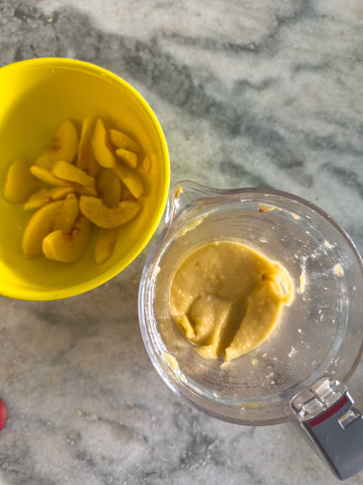 A yellow bowl with sliced peaches next to a blender containing thick peach puree, all on a gray marble countertop.