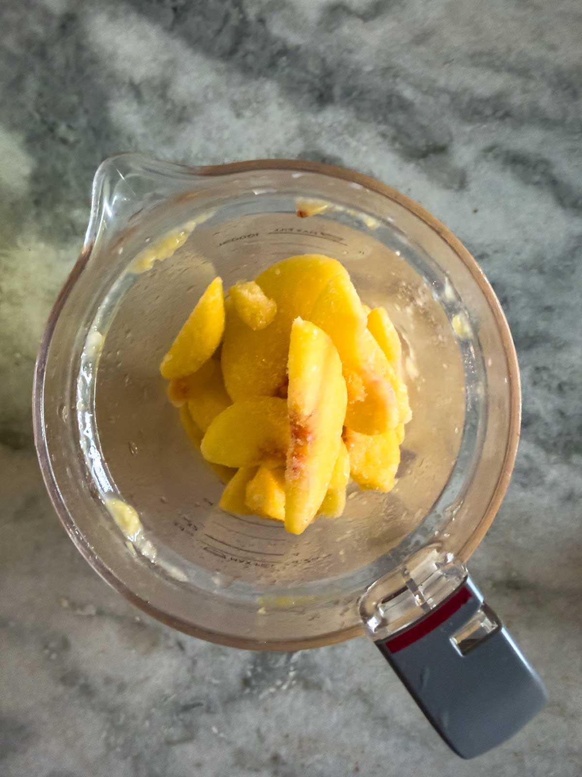 A glass blender pitcher filled with frozen peach slices sits on a gray marble countertop, viewed from above.