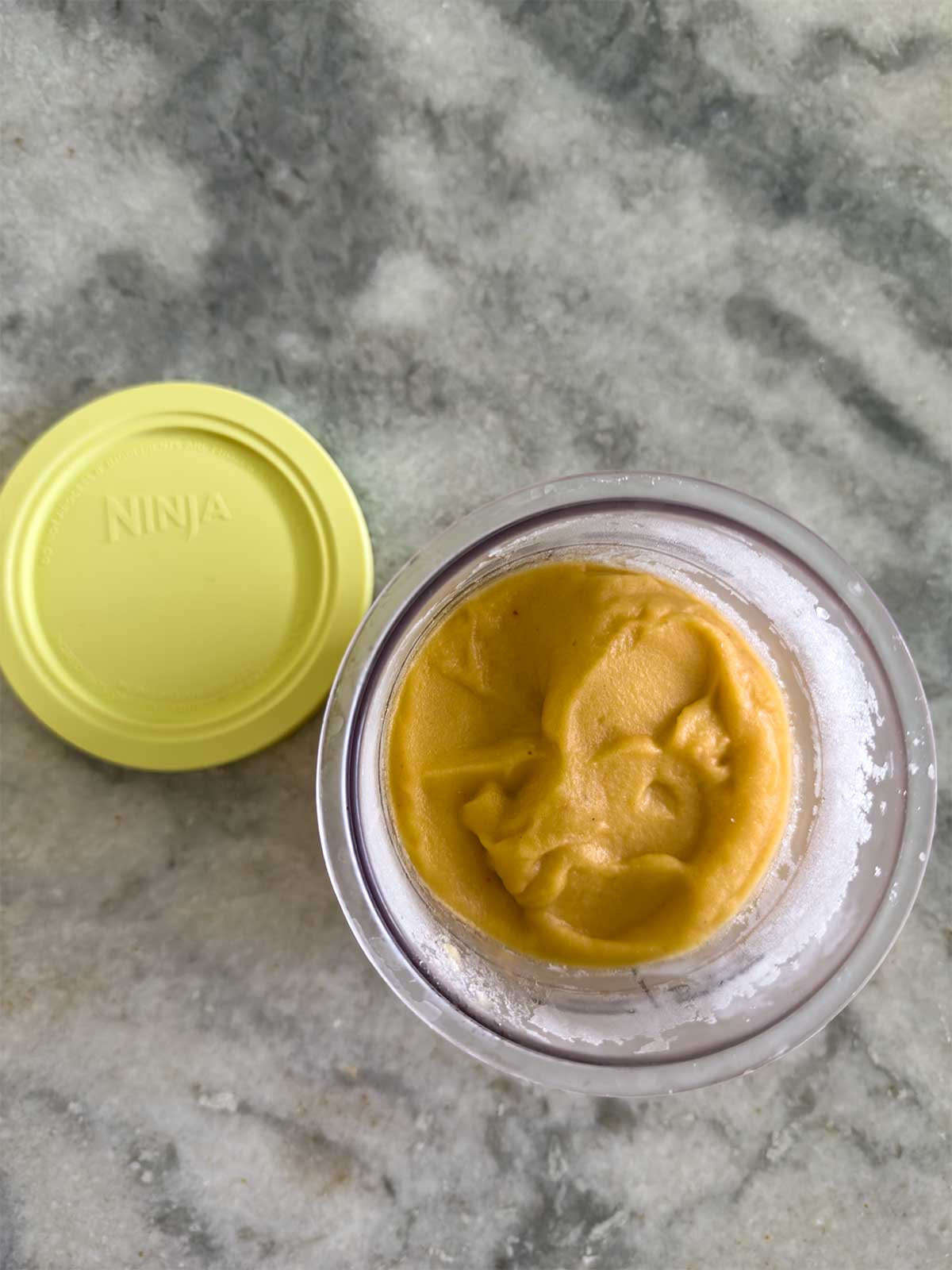A glass container with a yellow lid labeled “Ninja” sits on a marble surface, filled with a smooth, yellowish-orange frozen mixture. The lid is off and placed beside the container.