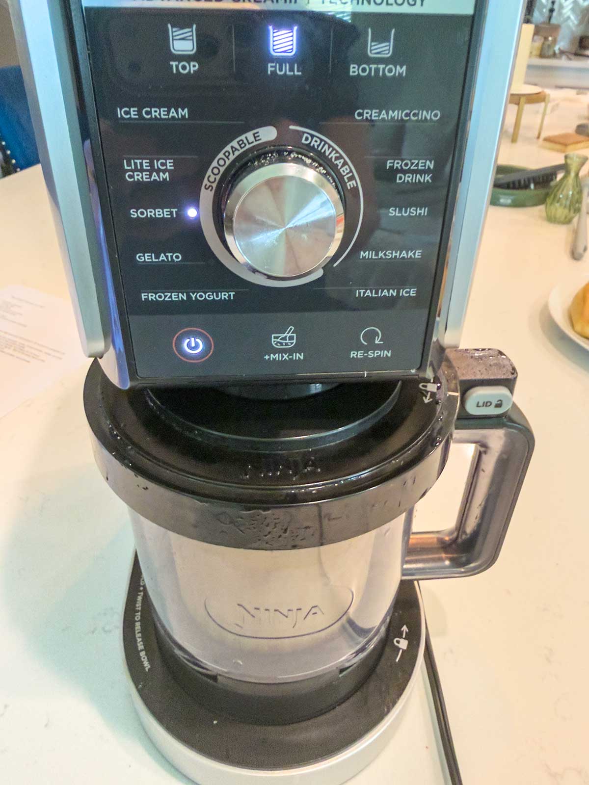 A Ninja ice cream maker on a counter, showing its control panel with settings for ice cream, sorbet, gelato, frozen yogurt, and other frozen treats. A large dial and several buttons are visible.