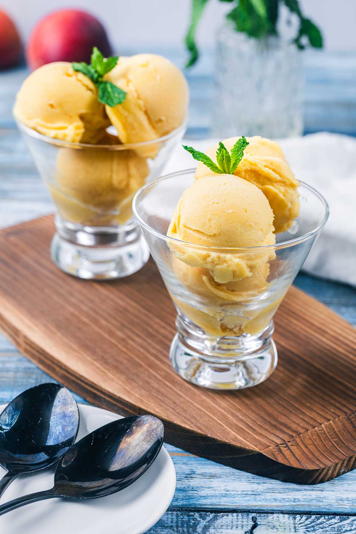 Two glass dessert cups filled with scoops of peach sorbet, each garnished with a sprig of mint, are placed on a wooden board. Two black spoons rest on a white plate beside the desserts.