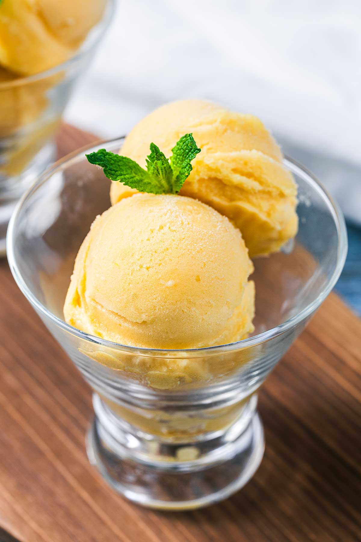 Two scoops of yellow mango sorbet in a clear glass, garnished with a fresh mint sprig, placed on a wooden surface with another glass of sorbet blurred in the background.