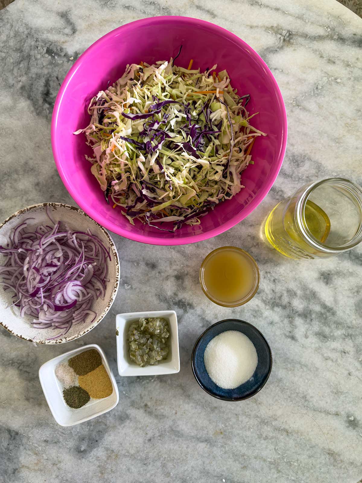 Ingredients for Old-Fashioned Coleslaw with Vinegar on a marble surface: shredded green and purple cabbage in a pink bowl, sliced red onions, capers, oil, vinegar, sugar, and spices arranged in small containers.
