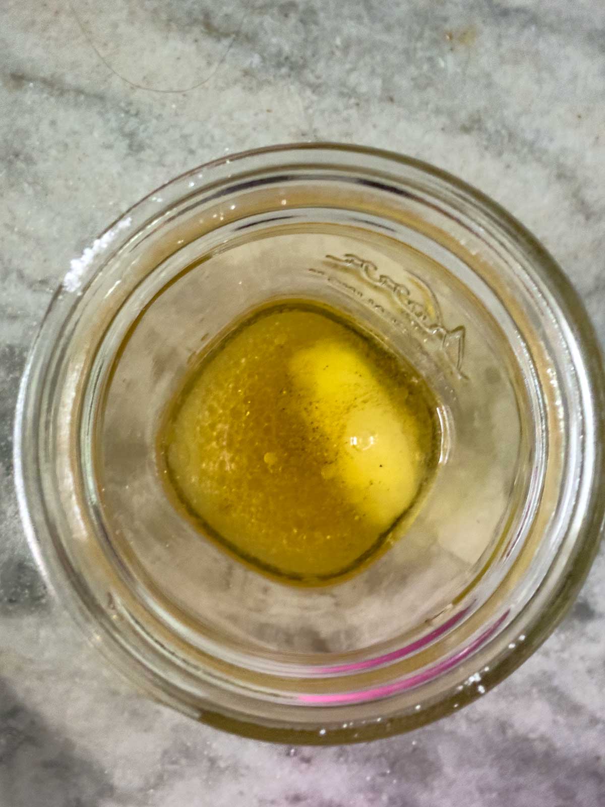 A top-down view of a glass jar containing a small amount of yellow liquid with some bubbles, possibly the tangy dressing for Old-Fashioned Coleslaw with Vinegar, sitting on a gray marble surface.