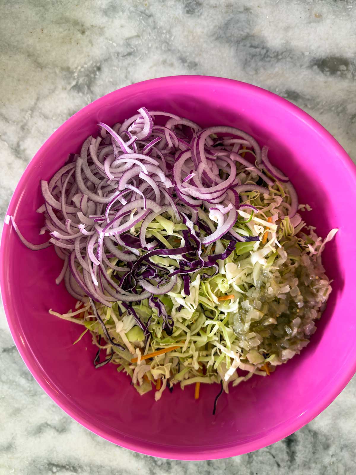 A pink bowl of Old-Fashioned Coleslaw with Vinegar, featuring thinly spiralized purple onions, shredded green and purple cabbage, and finely chopped pickles, sits on a marble countertop.