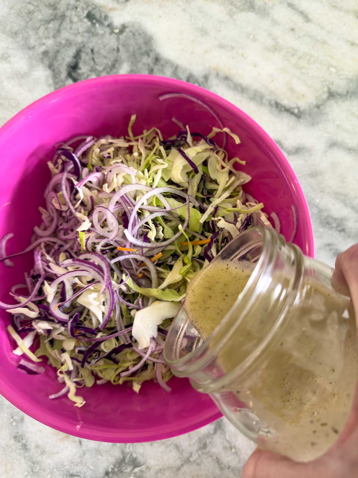 A hand pours creamy dressing from a jar onto shredded cabbage and carrots in a bright pink bowl, creating an Old-Fashioned Coleslaw with Vinegar on a marble countertop.