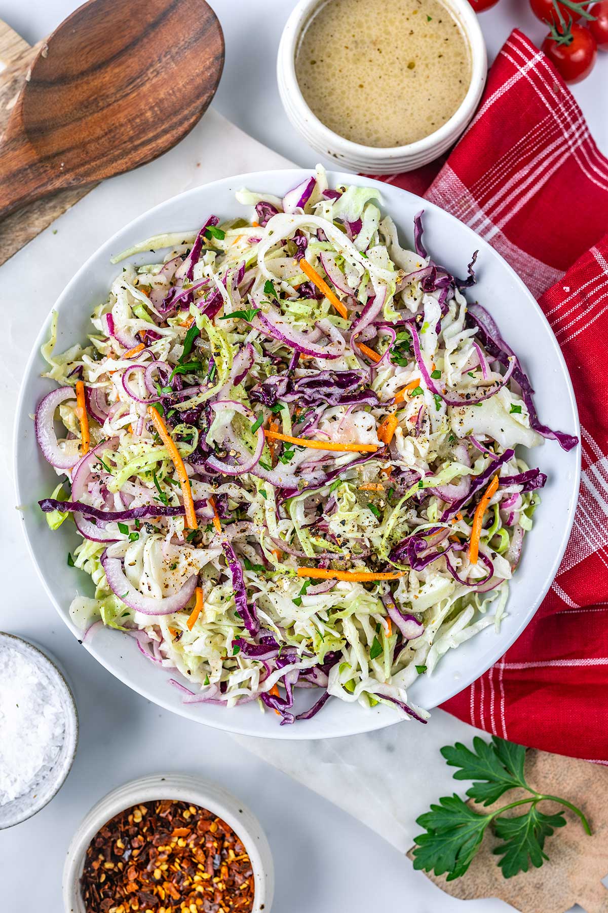 A bowl of coleslaw with shredded green and purple cabbage, carrots, and red onion, topped with black pepper. Surrounding the bowl are a bowl of dressing, sautéed mushrooms for steak, a red napkin, seasonings, and a wooden spoon.