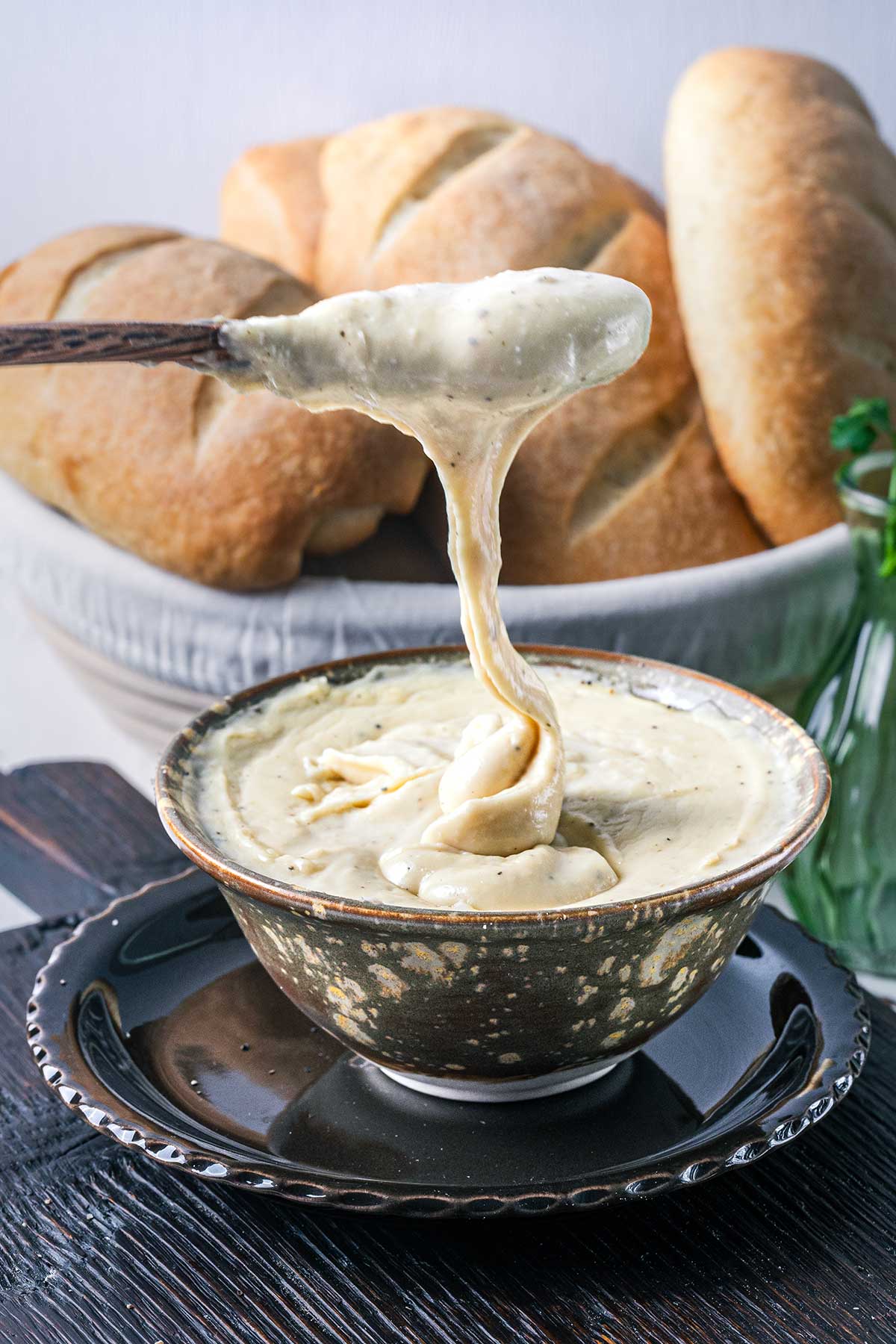 A spoon lifts a thick, creamy cheese dip from a rustic bowl, with fresh crusty bread loaves in the background. The cheese stretches from the bowl to the spoon.