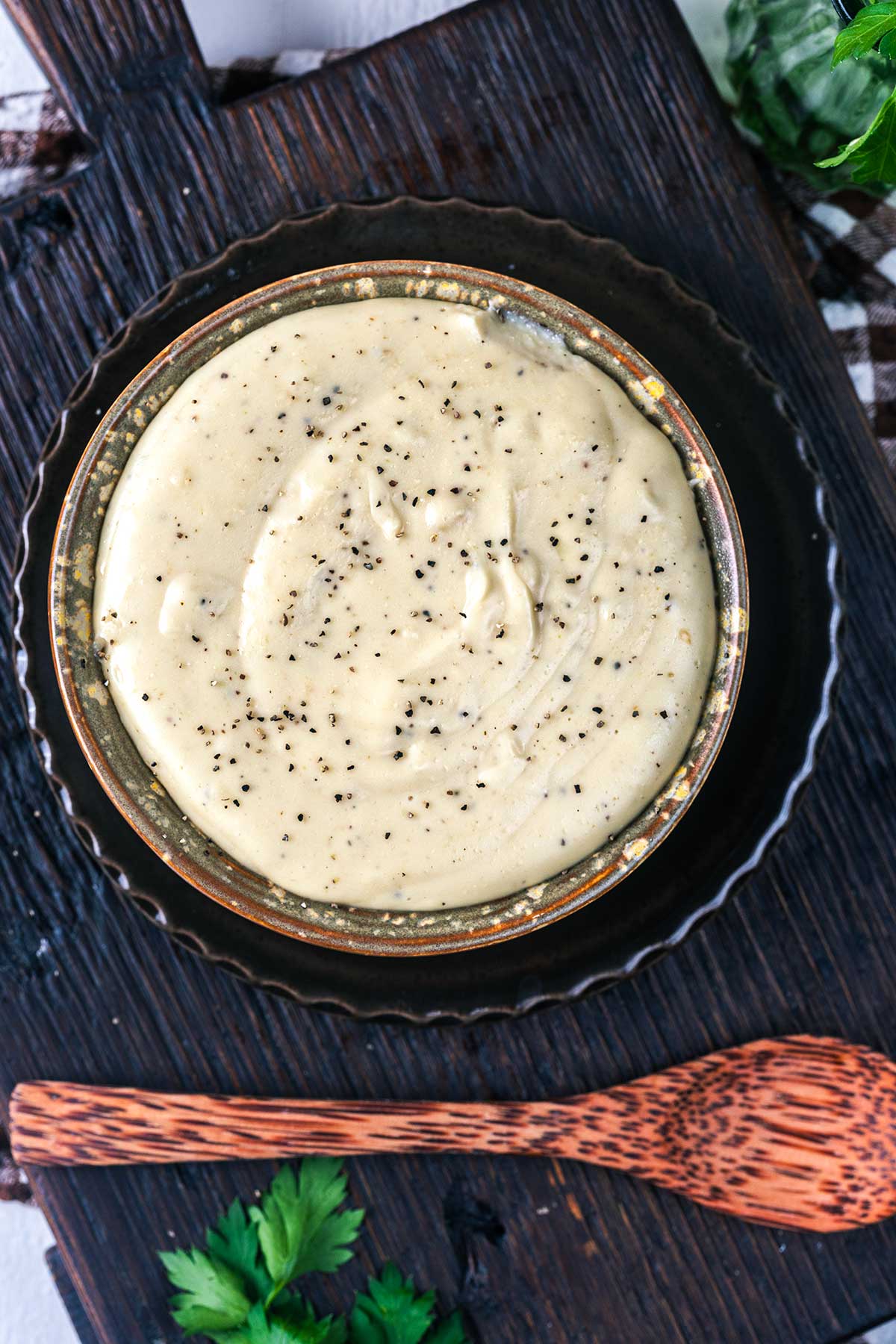 A bowl of creamy dip sprinkled with black pepper sits on a dark wooden tray next to a patterned wooden spoon and some fresh parsley.
