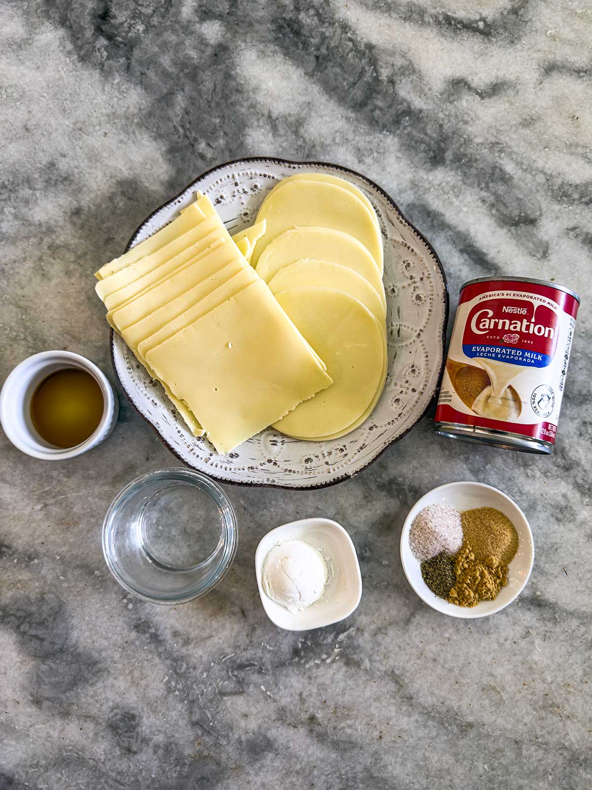 A plate with slices of cheese, a can of evaporated milk, a small bowl of liquid, an empty glass bowl, and two small bowls containing various powdered spices on a marble countertop.