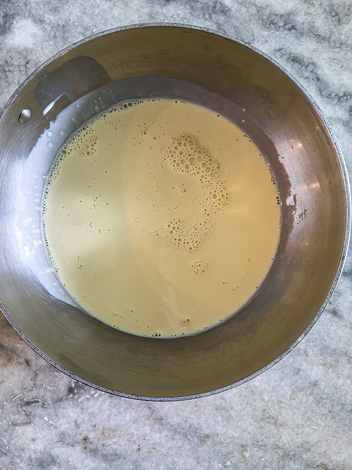 A metal mixing bowl filled with a pale yellow liquid batter sits on a gray marble countertop.