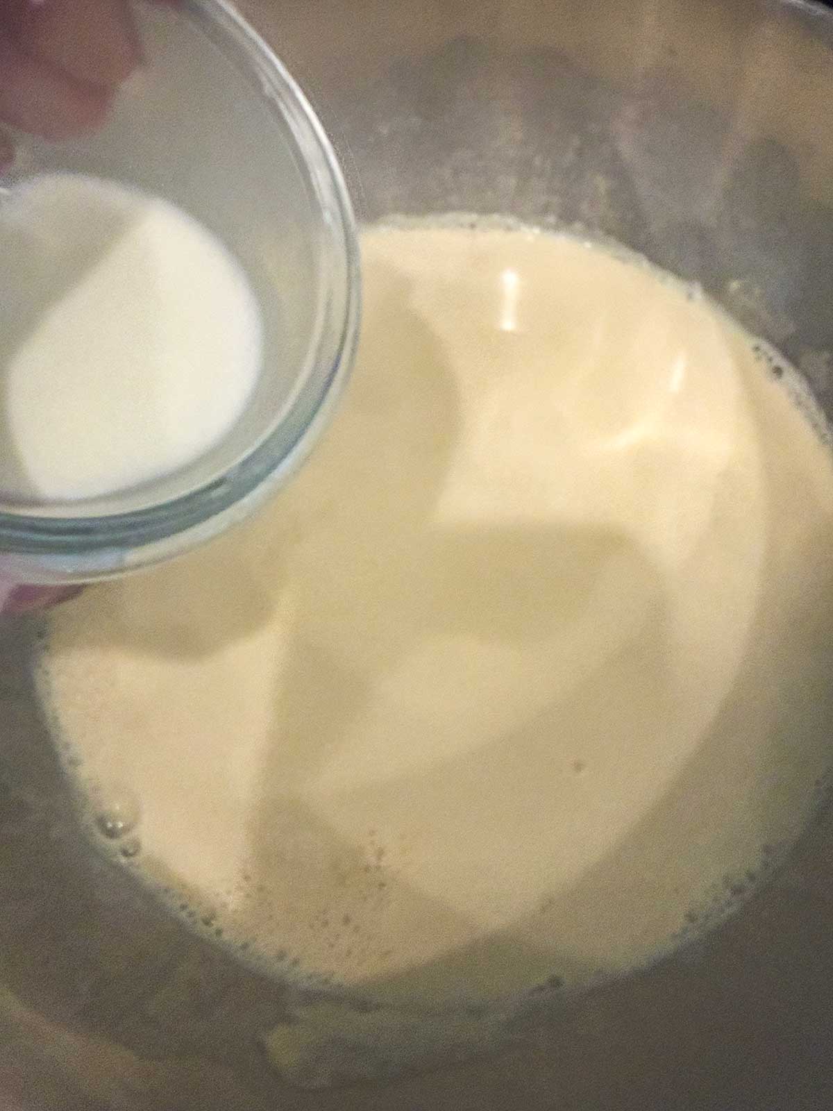 A hand holds a small glass bowl of milk above a larger bowl containing a tan-colored liquid mixture, suggesting an ingredient is about to be added while cooking or baking.