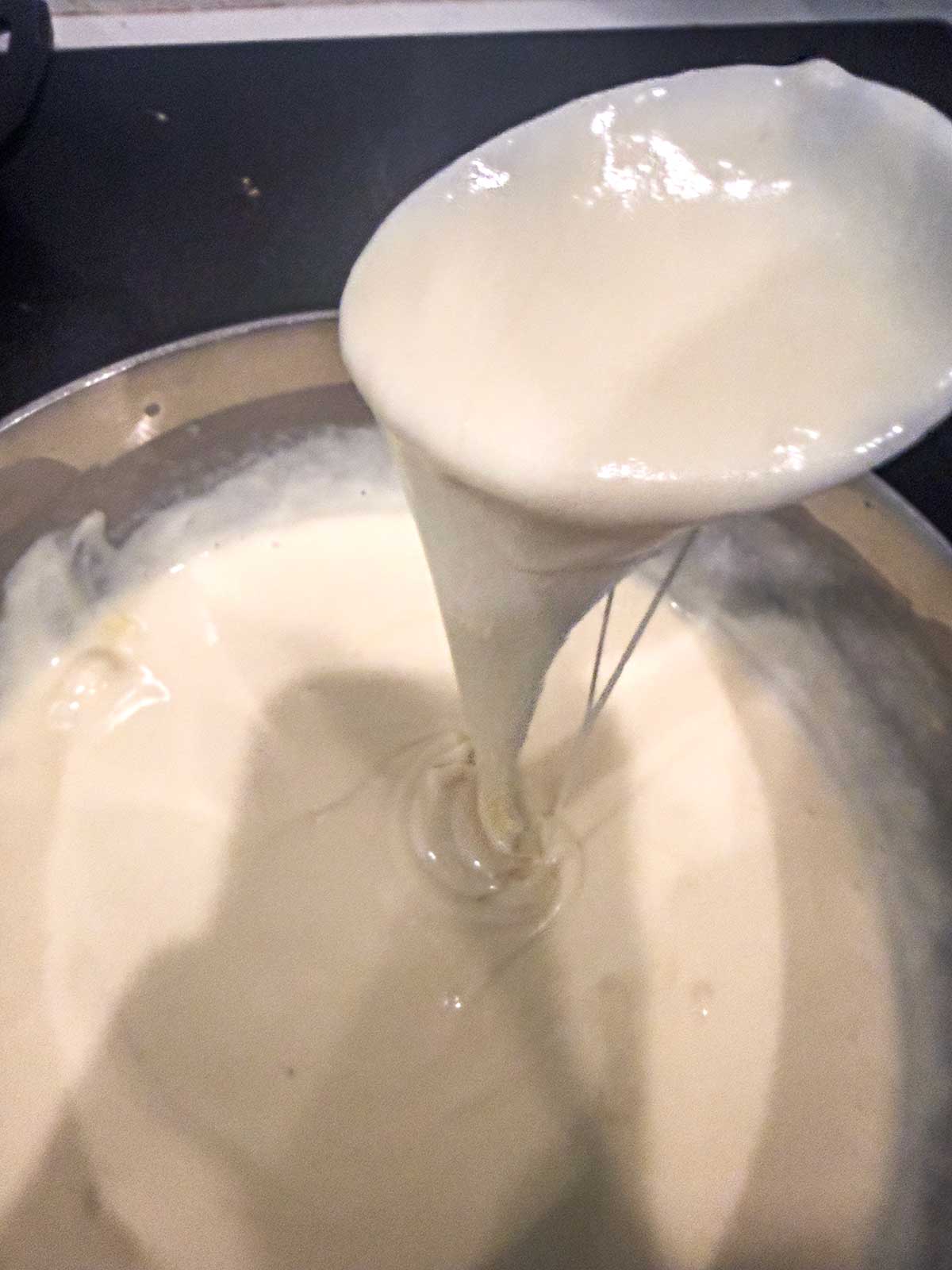 A close-up of a spoon lifting thick, creamy white sauce from a pot, showing the smooth, glossy texture as it pours slowly back into the pot.