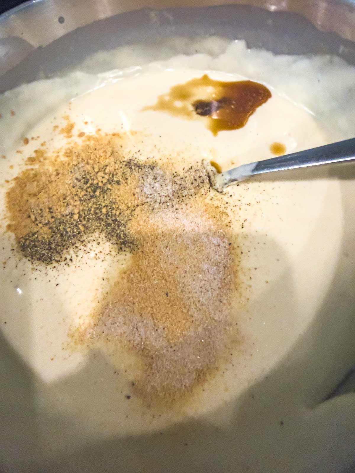 A close-up of a creamy sauce in a bowl with various spices, pepper, and a liquid seasoning on top, being stirred with a metal spoon.