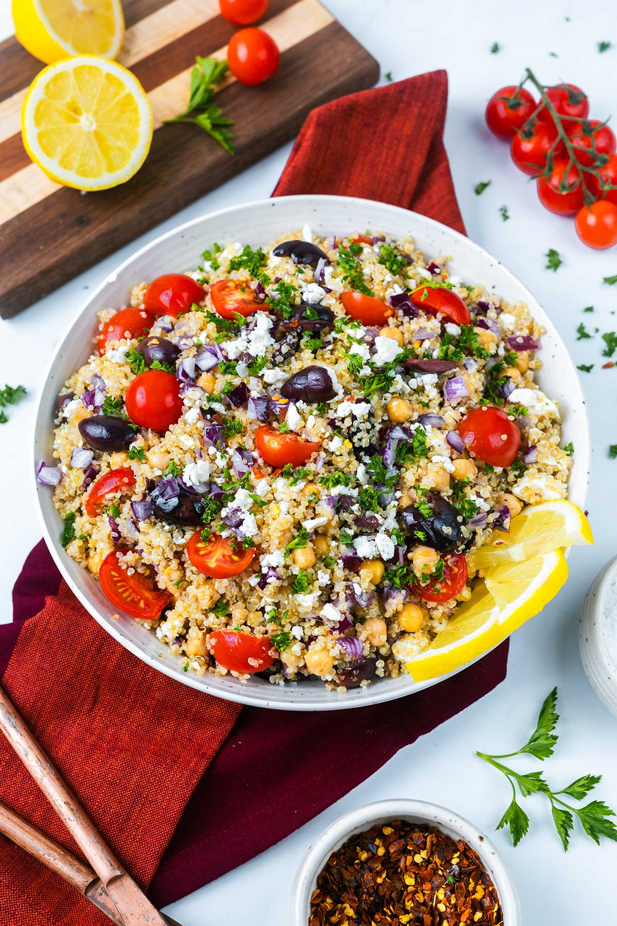 A bowl of Mediterranean quinoa salad with cherry tomatoes, black olives, chickpeas, red onions, parsley, and feta, garnished with lemon slices. Cutting board with lemons and tomatoes in the background.