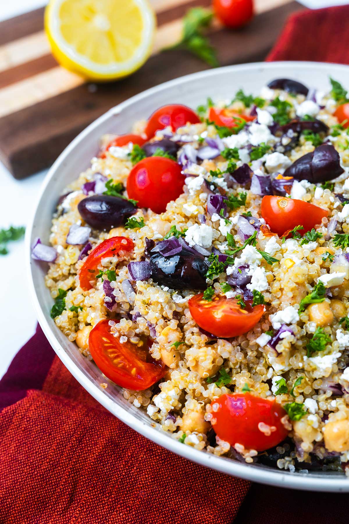 A bowl of quinoa salad topped with cherry tomatoes, black olives, crumbled feta cheese, chopped red onion, and parsley. A sliced lemon and tomatoes are in the background on a wooden cutting board.