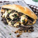 A sandwich filled with grilled steak slices, sautéed onions, and creamy cheese sauce, garnished with chopped parsley, served on a rustic bread roll on a marble surface.