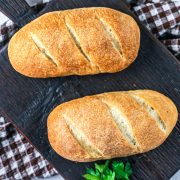 Two small loaves of golden-brown bread with three diagonal slashes on top, placed on a dark wooden surface, with a checkered cloth and some green herbs nearby.