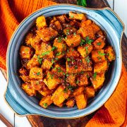 A bowl filled with glazed, cubed sweet potatoes garnished with chopped parsley, set on an orange napkin. The sweet potatoes appear caramelized and seasoned, giving them a rich, appetizing color.