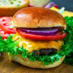 A close-up of a cheeseburger with cheddar cheese, lettuce, tomato slices, red onion rings, and a shiny brioche bun, set on a wooden surface with leafy greens and tomato slices in the background.