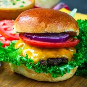 A close-up of a cheeseburger with cheddar cheese, lettuce, tomato slices, red onion rings, and a shiny brioche bun, set on a wooden surface with leafy greens and tomato slices in the background.