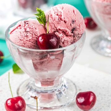Two scoops of pink cherry ice cream in a glass dessert dish, garnished with a fresh cherry and a sprig of mint, with additional cherries placed around the dish on a white surface.