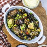 A white oval dish filled with crispy roasted Brussels sprouts, sprinkled with grated cheese and spices, sits on a wooden board next to a small bowl of grated cheese and a brown checkered cloth.