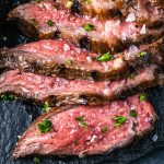 Close-up of sliced grilled sirloin steak on a dark surface, topped with coarse salt and chopped fresh herbs. The meat appears juicy and tender, with a pink center and seared edges.