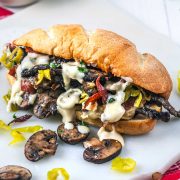 A toasted sandwich filled with grilled mushrooms, sliced pepperoni, meat, and topped with creamy white sauce, sits on a white surface with extra mushroom slices and pepper rings nearby.