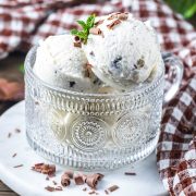 Two scoops of vanilla ice cream with chocolate flakes in a decorative glass cup, garnished with a mint sprig and chocolate shavings, placed on a white surface with a brown and white checkered cloth in the background.