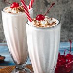 Two tall glasses of vanilla milkshake topped with whipped cream, chocolate shavings, and a cherry, each with a red-and-white striped straw. A bowl of cherries is visible in the background.